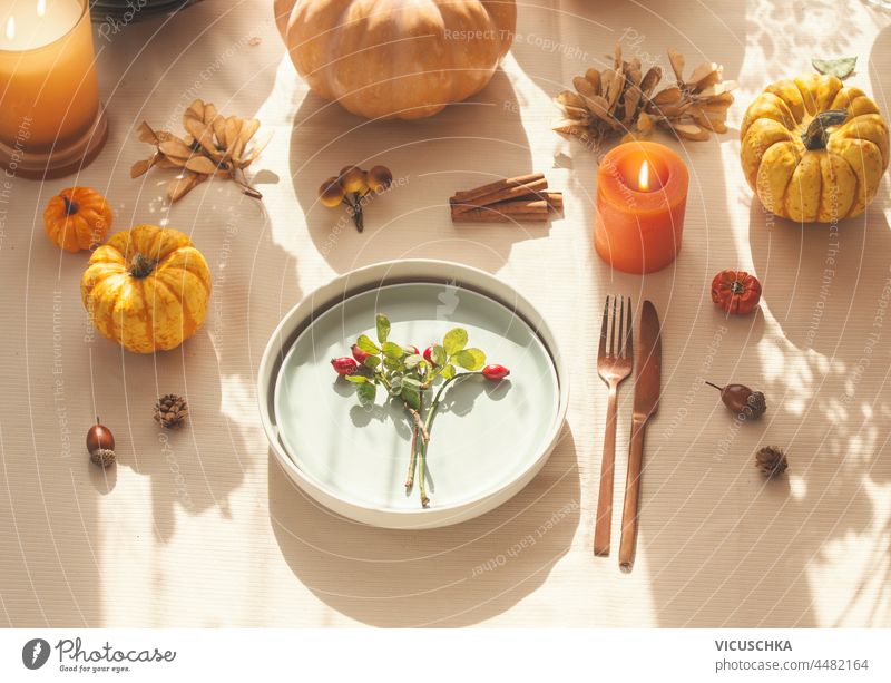 Autumn table setting with pumpkins, burning candles, acorn and plate with rose hip and cutlery on table with beige tablecloth. Cozy, sunny autumn concept with natural light. Top view.