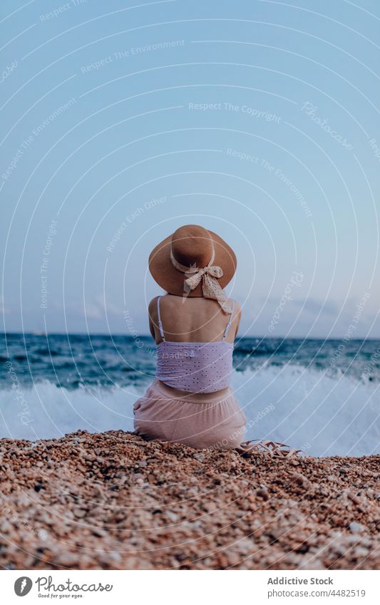 Anonymous woman in hat sitting near sea coast rock summer nature water beach outfit sandal female rocky stone shore environment admire ripple peaceful seaside