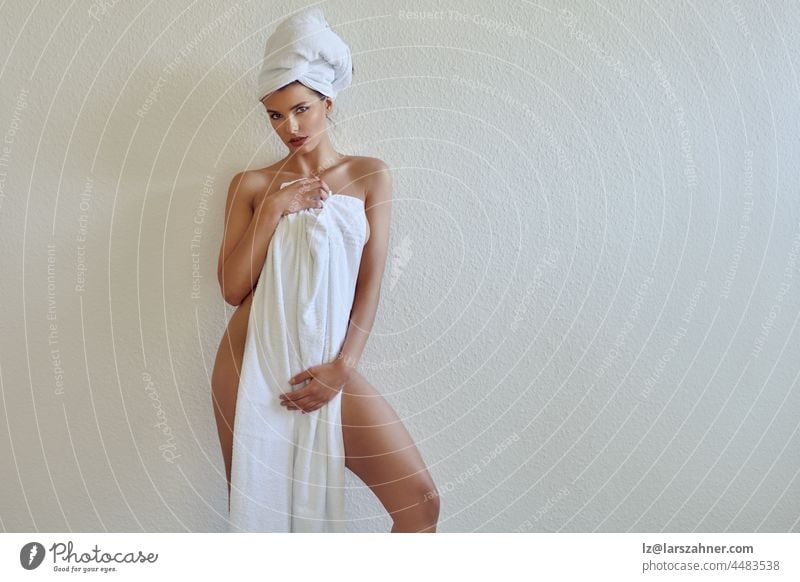 Sexy beautiful young woman with her hair twisted up in a towel holding a second white towel draped to her chest as she gives the camera a sultry look in a three-quarter pose against a wall with copy space