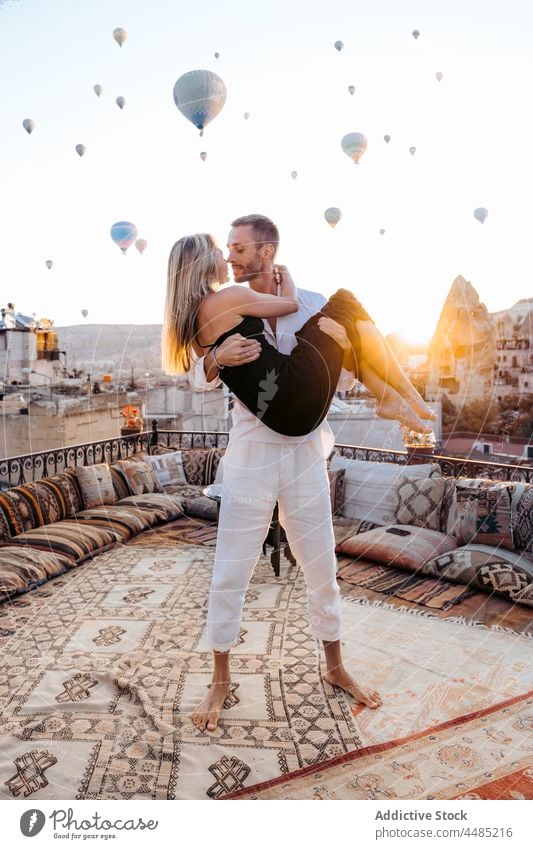Male holding soulmate on terrace against hot air balloons couple embrace rooftop romantic together love affection relationship date bonding weekend pleasure