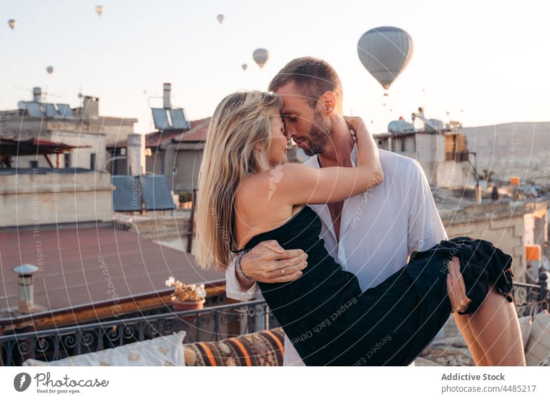 Male holding soulmate on terrace against hot air balloons couple embrace rooftop romantic together love affection relationship date bonding weekend pleasure