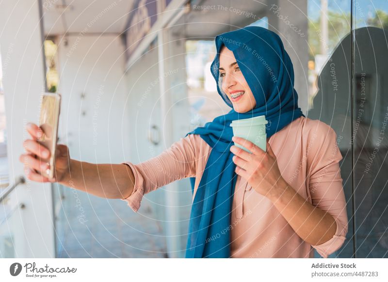 Smiling Muslim woman taking selfie in city smartphone hijab takeaway coffee self portrait smile tradition female arab ethnic muslim street headscarf happy using