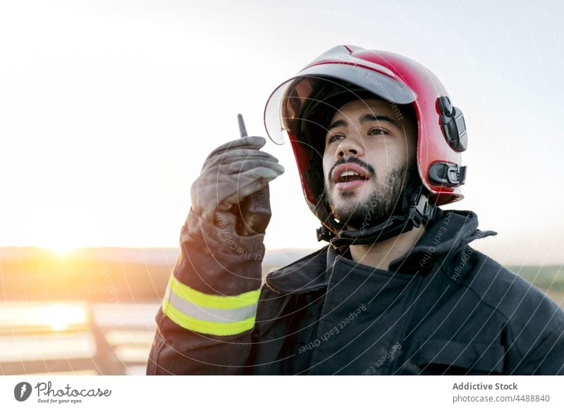 Young fireman speaking into transmitter firefighter helmet uniform report walkie talkie professional occupation job career glove positive male safety confident
