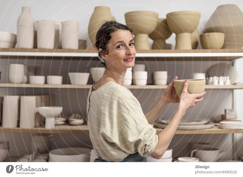 Young female ceramist indoors holding handmade clay product. Conception of pottery craft woman artisan smiling looking at camera hand-made workshop business
