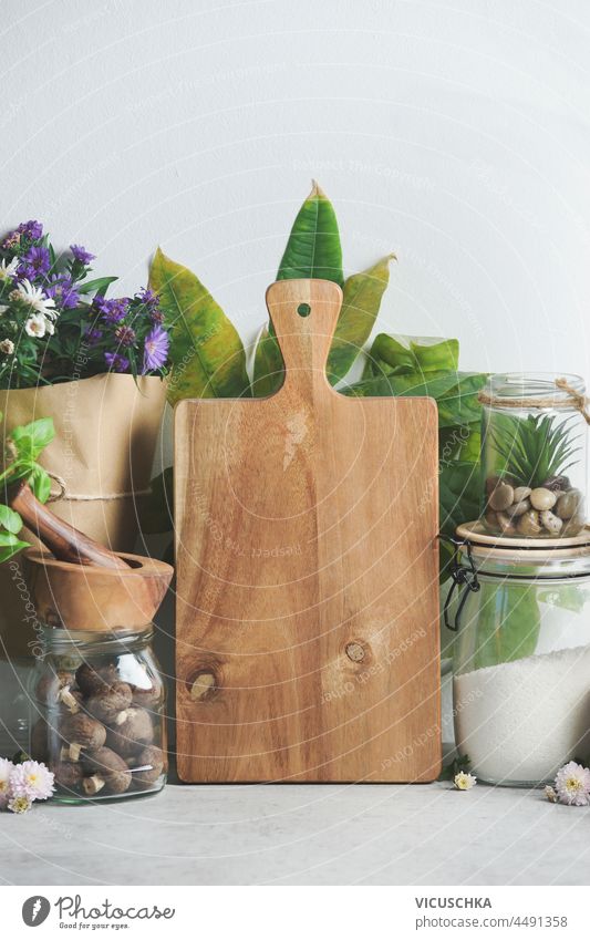 Sustainable kitchen concept with  plastic free packaging. Wooden cutting board, glass jar with flour and potted flowers at white wall background. Modern lifestyle. Front view with copy space.