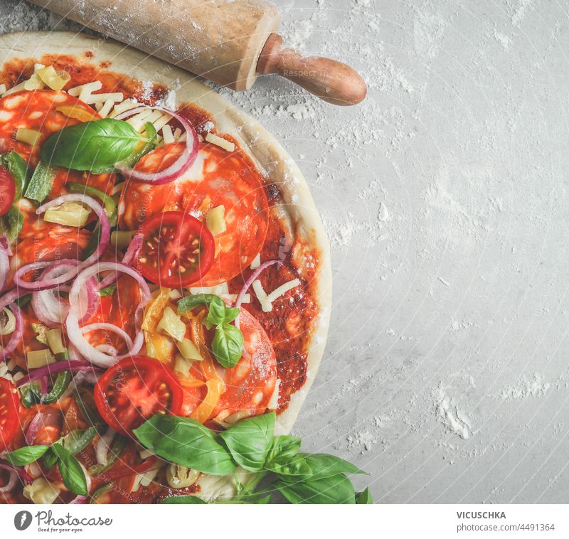 Close up of fresh pizza with tomatoes, onion, basil, cheese and salami and rolling pin on grey table with flour. Cooking traditional italian food at home with fresh ingredients. Top view. Copy space.
