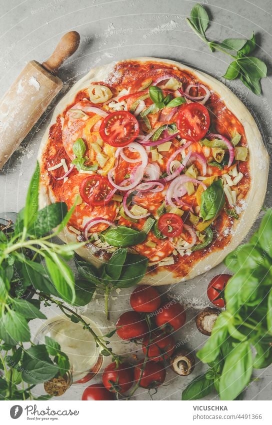 Pizza preparation with salami, tomatoes, mushrooms, onion, basil, cheese , fresh herbs and rolling pin on grey table. Cooking traditional Italian food at home with fresh ingredients. Top view