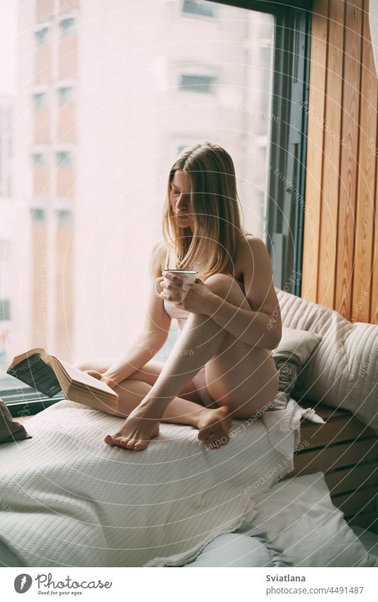 A young girl is sitting on the windowsill in the early morning, reading a book and drinking hot coffee woman leisure home cup people house indoor interior