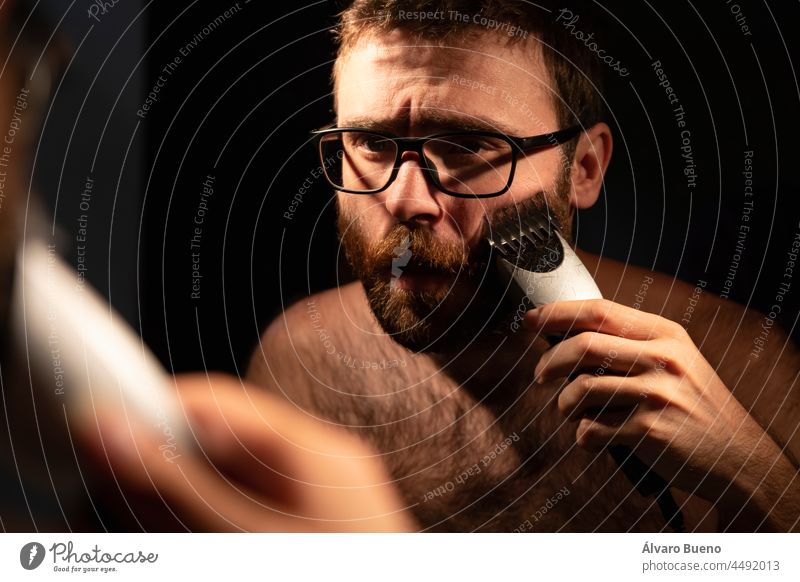 A young man shaving and trimming his beard with an electric razor, in the bathroom at home, early in the morning, to be groomed before going out