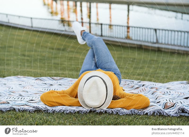back view of woman lying on grass wearing hat and yellow pullover. Crossed legs. Relax outdoors in park with lake. Lifestyle relax madrid city sleeping
