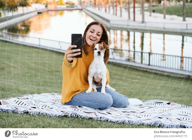 young caucasian woman Woman taking picture with mobile phone. woman having fun with jack russell dog in park, sitting on blanket during autumn season. Pets and love concept
