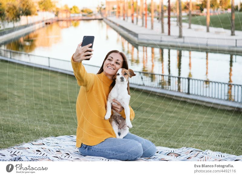 woman having fun with jack russell dog in park, sitting on blanket during autumn season. Woman taking picture with mobile phone. Pets and love concept happy