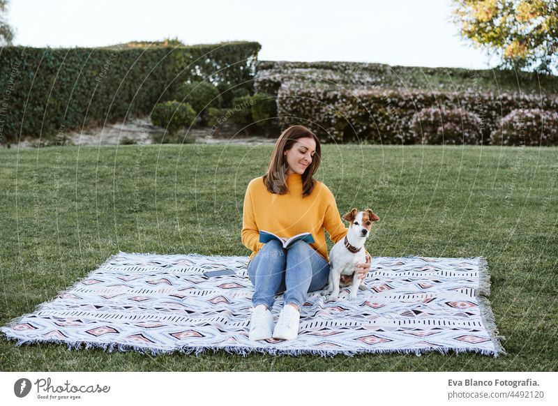 happy woman having fun with jack russell dog in park, sitting on blanket during autumn season. Woman reading a book while hugging dog. Pets and love concept