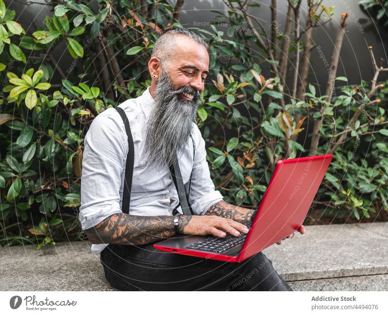 Cheerful bearded businessman typing on laptop on street hipster online internet freelance plant work style modern trendy confident tattoo outfit male fashion