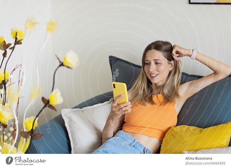 Happy young caucasian blonde woman relaxing at home alone, sitting on couch in comfortable pose, blogger sharing good news at social media via mobile phone. Smiling woman enjoying weekend order goods food online in app