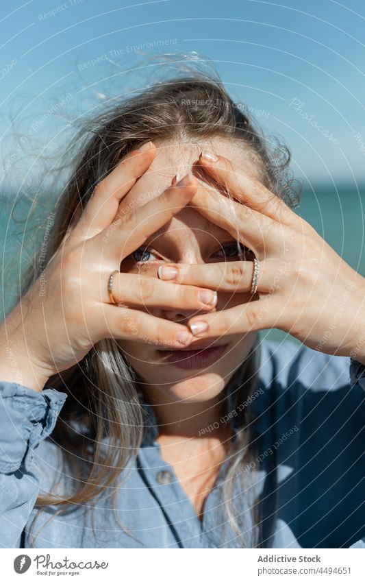 Woman looking through hands at camera woman summer sunlight coastline seashore trendy daytime wind hide peaceful appearance contemporary female gesture finger
