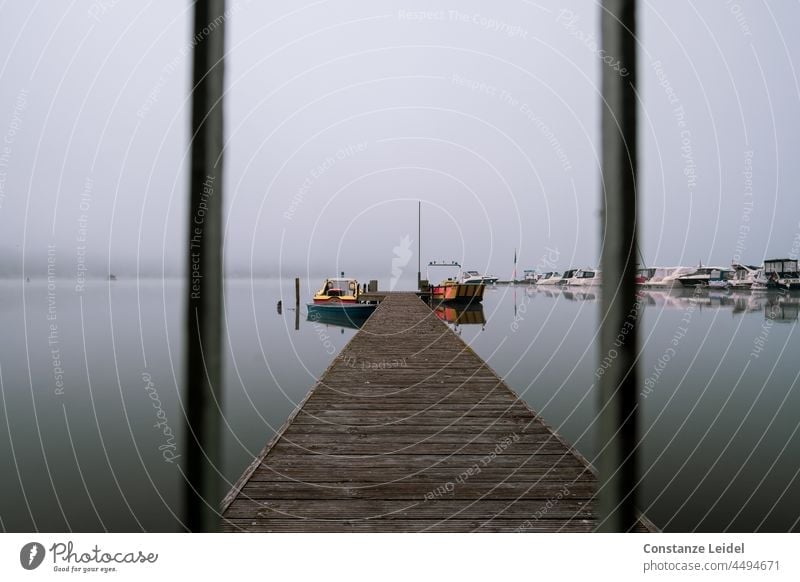 Wooden jetty with boats in the fog through a grate. Lake Water Calm Footbridge Dawn Morning Landscape Lakeside Fog Sky Colour photo Exterior shot Reflection