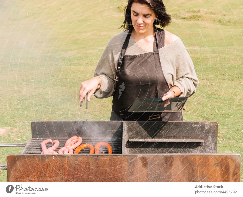 Woman roasting sausages on grill in countryside woman cook meat barbecue grate field bbq food tasty summer chef delicious picnic hot various assorted tong