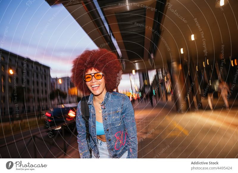 Cheerful woman walking on dark street afro style fashion trendy apparel outfit building evening twilight carefree house appearance female hairstyle sunglasses