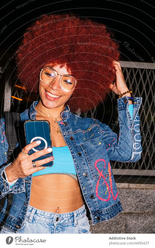 Positive woman browsing smartphone on dark street online afro evening urban fashion style trendy apparel surfing text message cellphone female gadget device