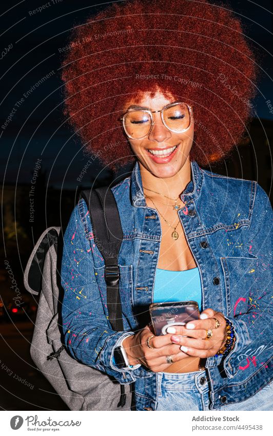 Positive woman browsing smartphone on dark street online afro evening urban fashion style trendy apparel surfing text message cellphone female gadget device