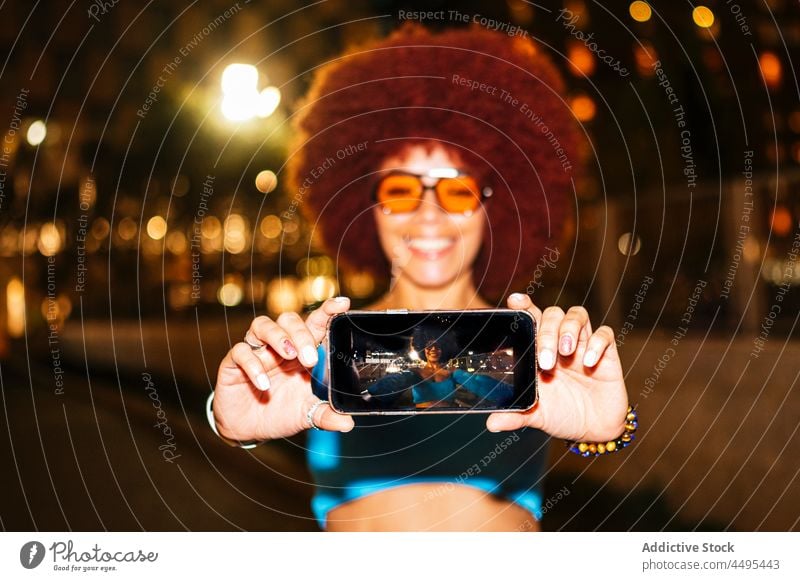 Cheerful woman taking selfie on dark street smartphone self portrait screen capture afro evening fashion urban photography memory style trendy apparel cellphone