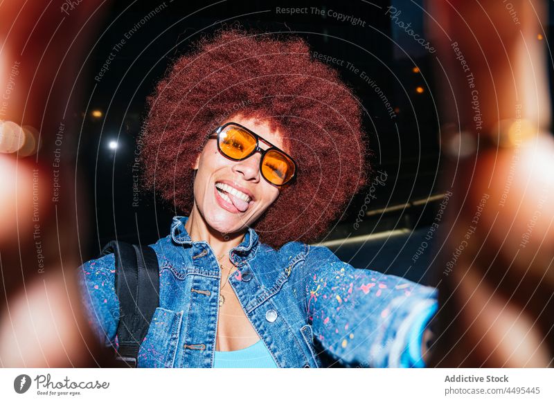 Cheerful stylish woman taking selfie on dark street self portrait capture memory grimace afro evening style fashion trendy apparel tongue out optimist female