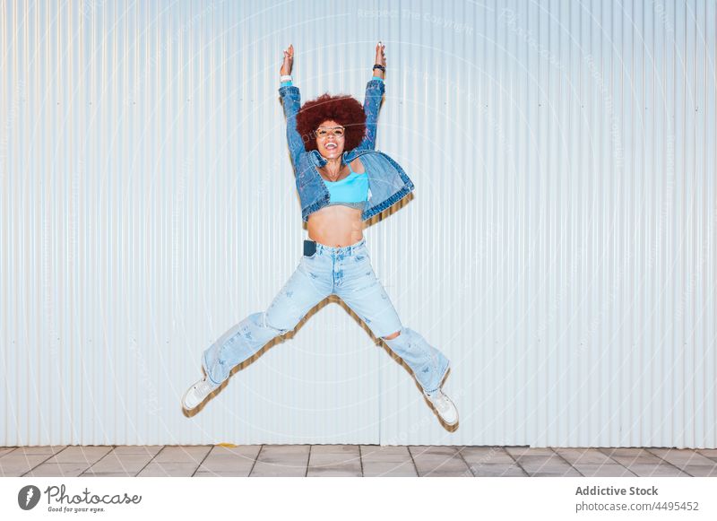 Happy woman jumping on street afro style fashion apparel wall outfit having fun carefree activity active energy arms raised mood trendy female hairstyle