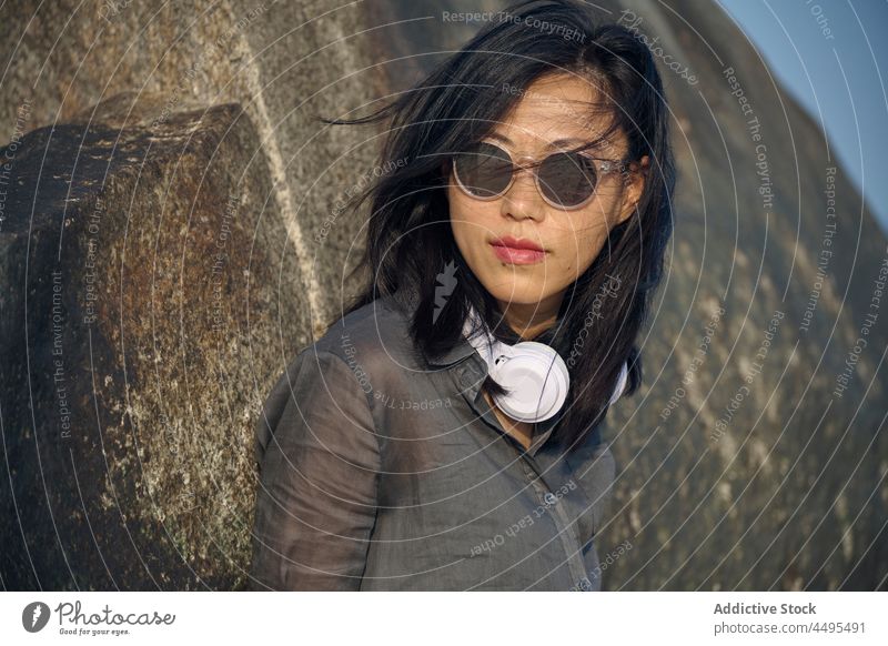 Stylish Asian woman in headphones standing near rock meloman stone formation nature style music mountain rocky flying hair female sunglasses trendy calm