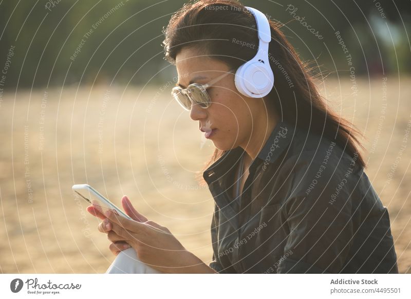 Asian woman browsing smartphone while listening to music on beach seaside seashore headphones meloman pastime online text message surfing internet cellphone