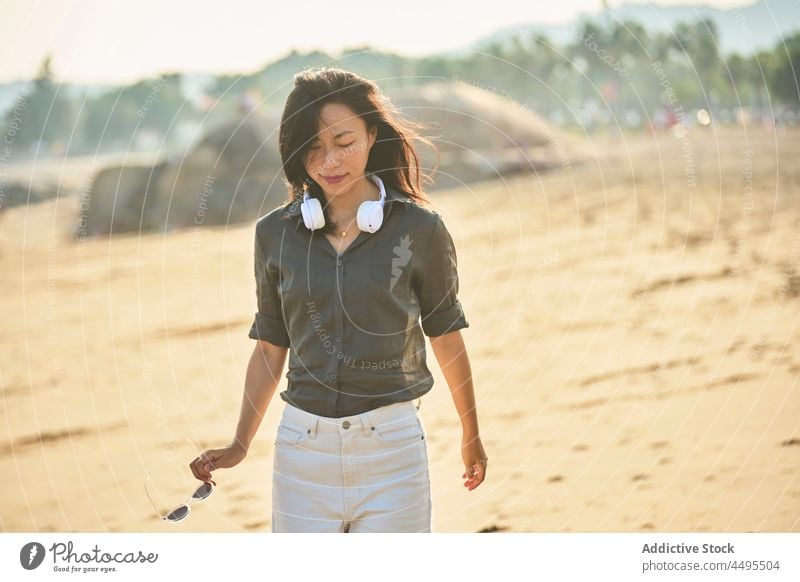 Pensive Asian woman listening to music on beach seaside seashore walk headphones meloman song pastime stroll thoughtful pensive playlist sand asian female