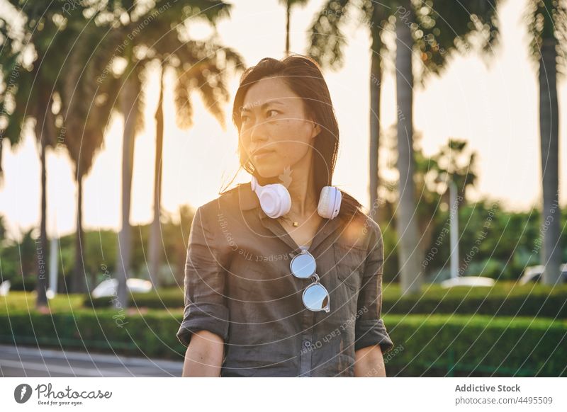 Asian woman with headphones on street meloman style music hobby road town pastime female trendy calm ethnic black hair attractive asian summer plant roadway