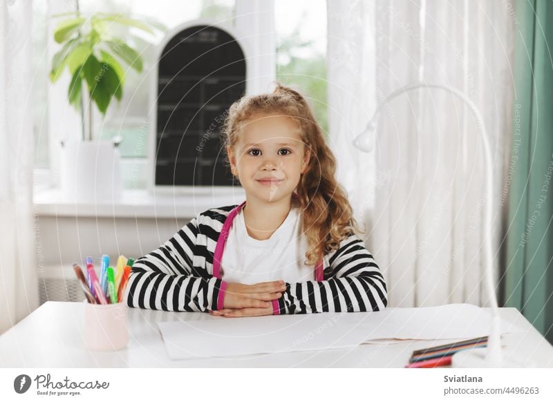 Portrait of a charming little girl sitting at a table. The girl is sitting at the table, on the table is a sketchbook, colored pencils. Homework, home training, social distance