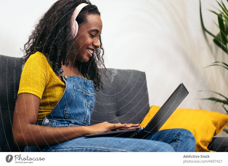 Happy African American woman using laptop in living room smile headphones music floor sofa delight denim home listen casual sit gadget cheerful browsing