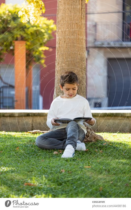Little kid reading a book in a shade of a tree boy calm caucasian child childhood concentrated day education exterior grass green happiness happy leaning