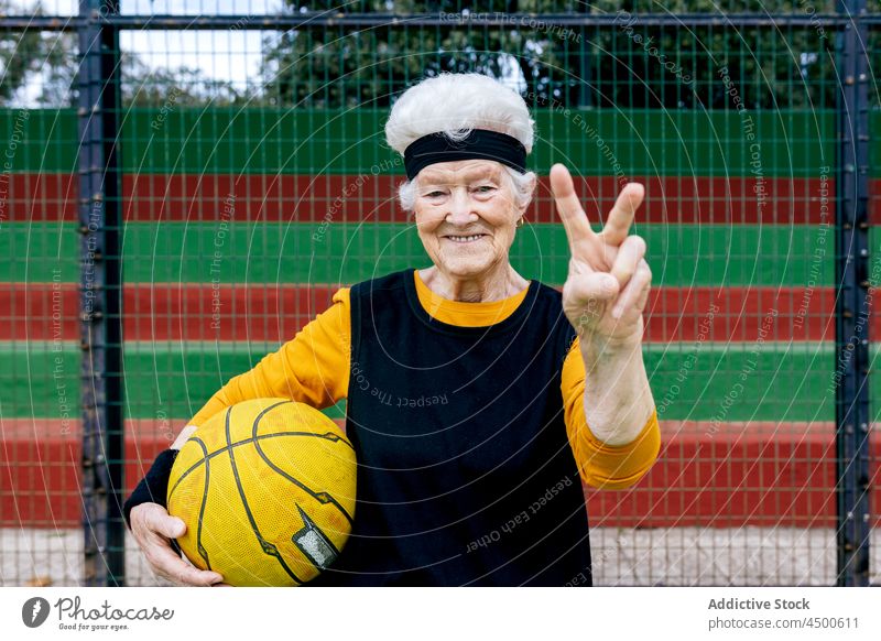 Positive senior woman with ball on playground basketball game headband sports ground two fingers sign peace victory healthy lifestyle hobby training mature