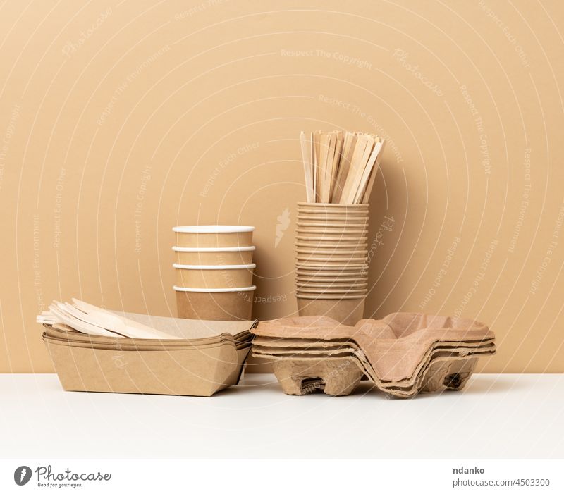 stack of brown disposable paper cups and a tray on a white table, wooden forks and knives, brown background americano beverage blank breakfast cafe caffeine