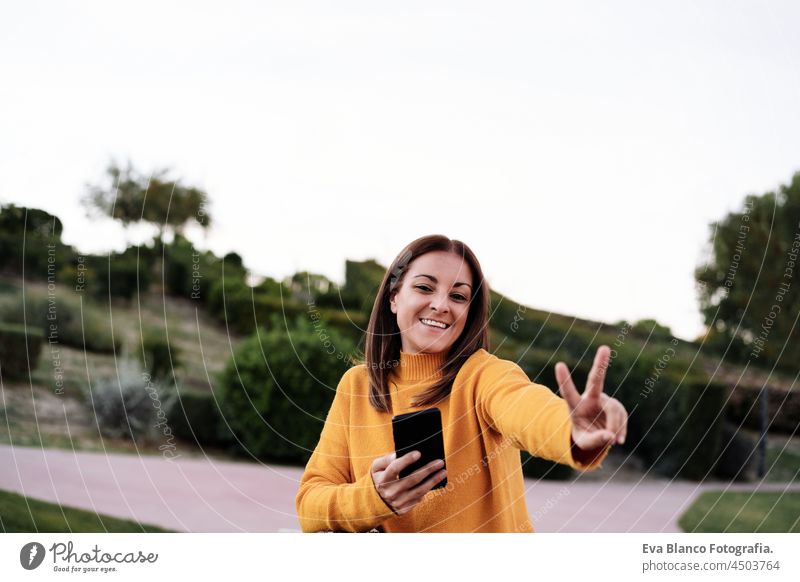 carefree caucasian woman making V sign wearing yellow pullover using mobile phone outdoors in park during sunset. Technology and lifestyle speaking technology