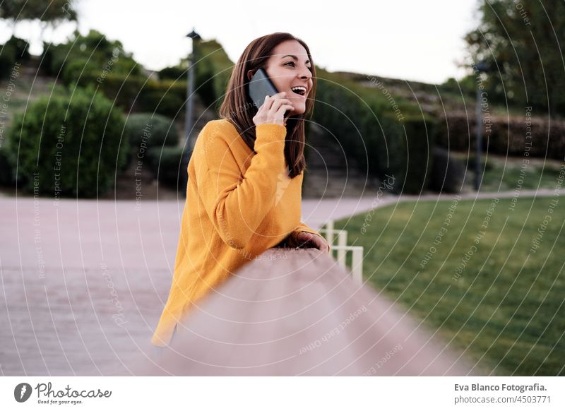 happy caucasian woman wearing yellow pullover talking on mobile phone outdoors in park during sunset. Technology and lifestyle speaking technology smart phone