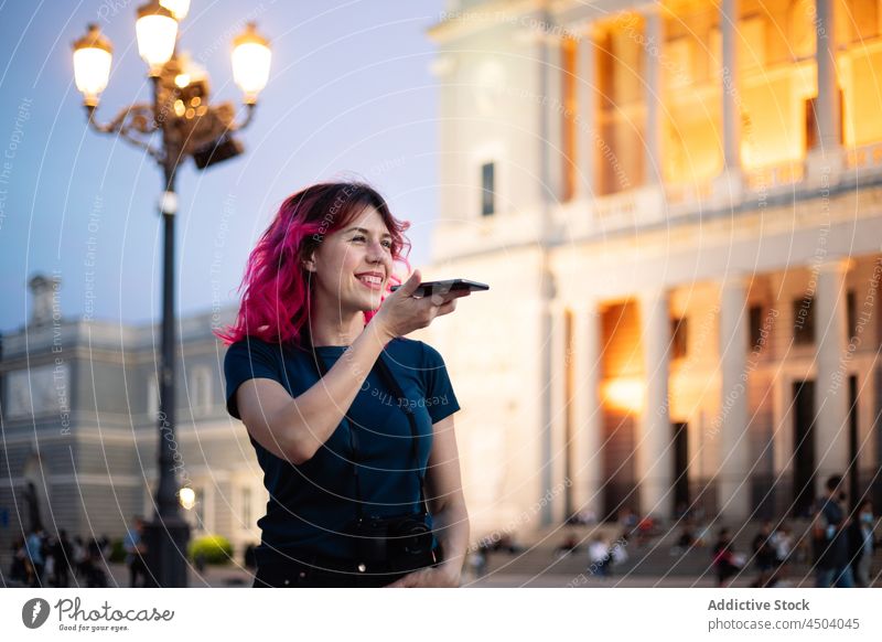 Positive woman recording audio message near historic building smartphone street voice aged classic internet online pink hair style female cellphone mobile