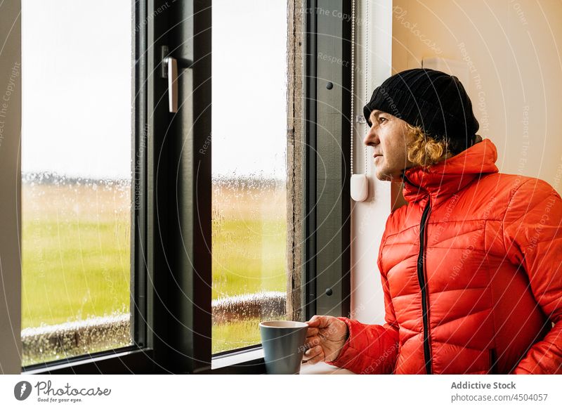Dreamy guy standing near window in kitchen and drinking coffee man dreamy traveler countryside hot drink recreation beverage breakfast rain male young