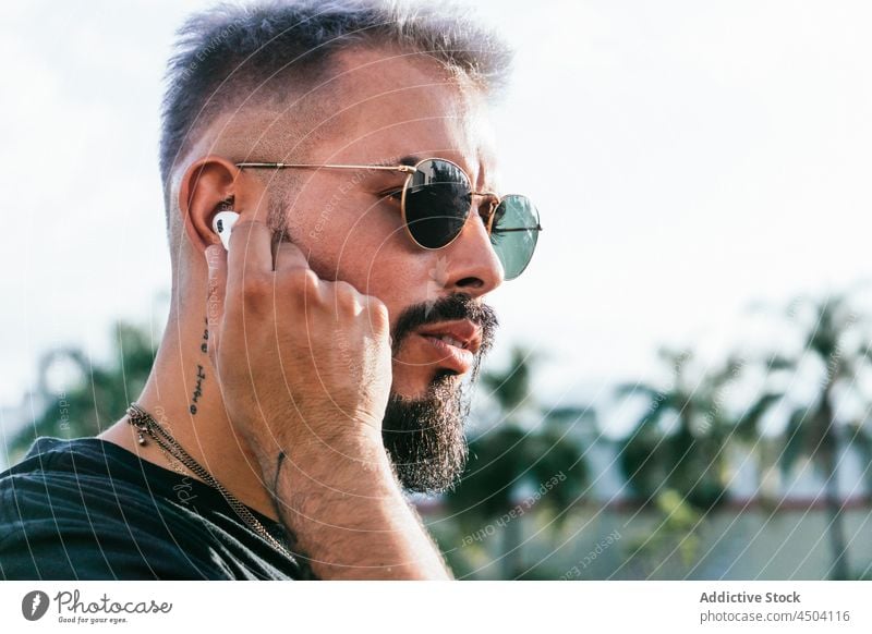 Serious male wearing earphones on street in daytime man tattoo sunglasses beard pensive trendy listen music thoughtful urban style appearance hairstyle green
