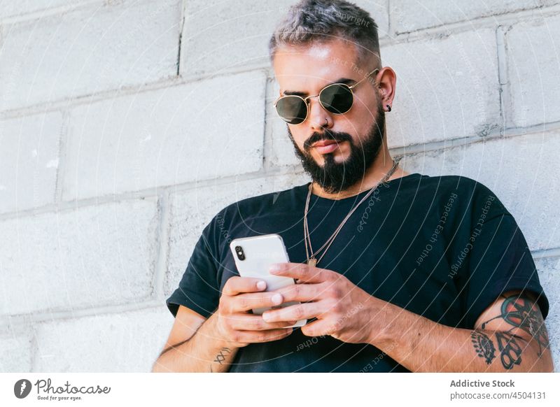 Cheerful male with tattoos using a smartphone man message mobile sunglasses gadget device street urban guy daylight communicate wall beard cellphone connection