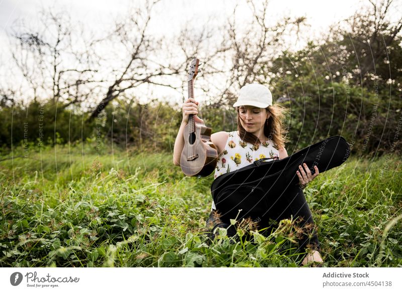 Woman musician opening case with ukulele on grass in daylight woman instrument hobby sit calm melody nature song sound talent thoughtful countryside green