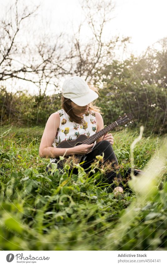 Female musician playing ukulele in nature in daytime woman instrument grass practice perform hobby female casual sit cap daylight calm style countryside talent
