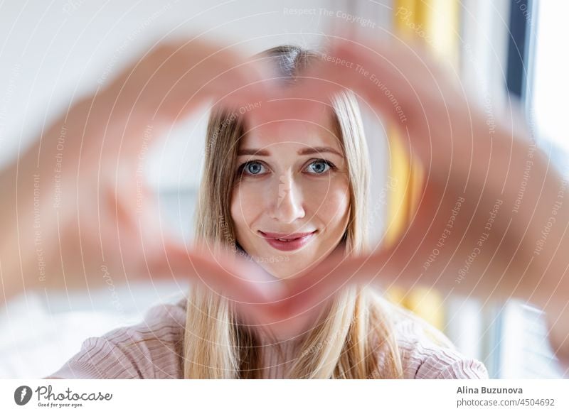 Close up smiling attractive young caucasian woman with blonde hair 30-35 years old showing heart shape gesture at home during coronavirus covid-19 pandemic quarantine and self isolation