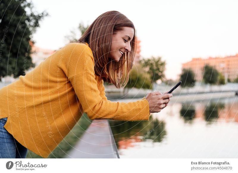smiling caucasian woman wearing yellow pullover using mobile phone device outdoors in park during sunset. Technology and lifestyle speaking technology