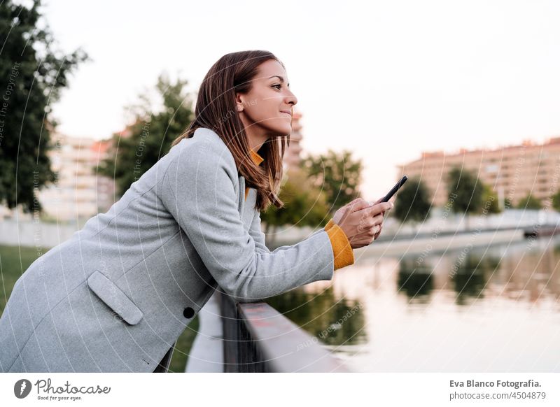 smiling caucasian woman wearing yellow pullover and grey coat using mobile phone device outdoors in park during sunset. Technology and lifestyle autumn speaking