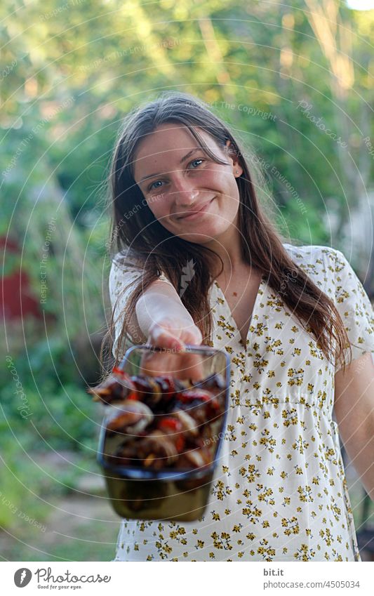Hospitality l who wants to have delicious skewers? I young woman in garden, wearing dress with flowers, smile on face; long open hair and bowl of glass with grilled food l grilling in nature, pure joy, happiness, warmth, vegetables warm, leaves green