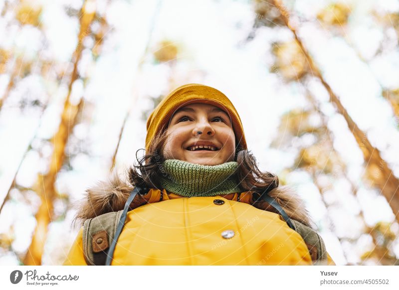Portrait of a cheerful caucasian tourist girl in an orange jacket and hat. Hiking in the autumn forest. Positive emotions. Outdoor activity. Hiking in the forest in the fall season. Close-up